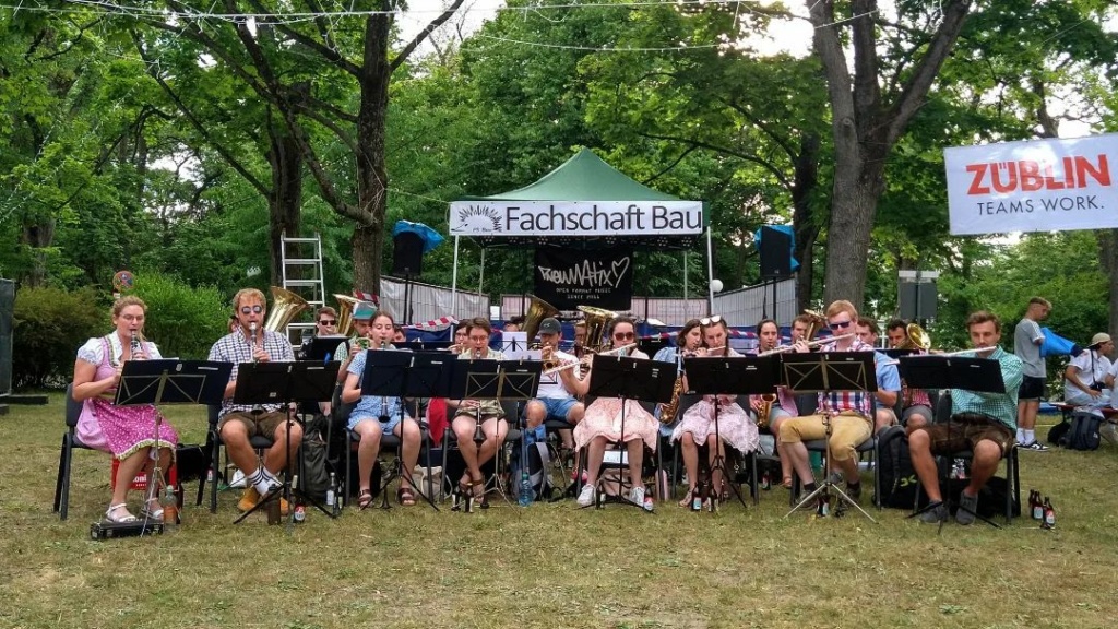 Musiker und Musikerinnen der KAmpus KApelle sitzen auf dem Rasen auf Stühlen und spielen ihre Blasinstrumente. Ein Banner mit der Aufschrift "Fachschaft Bau" hängt über ihnen.